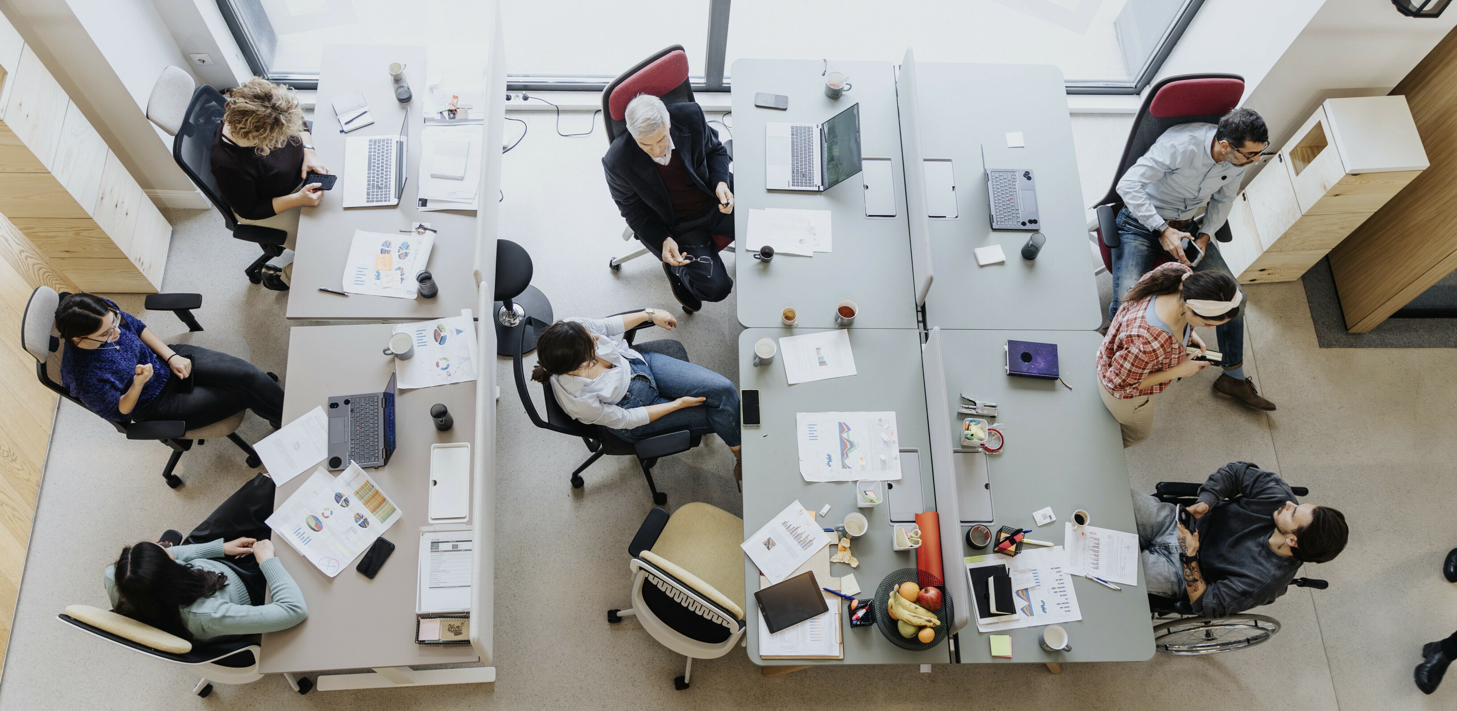 Overhead View Of A Collaborative Workspace With A Group Of People Engaged In Discussion, Working On Computers And Brainstorming Around A Shared Table.